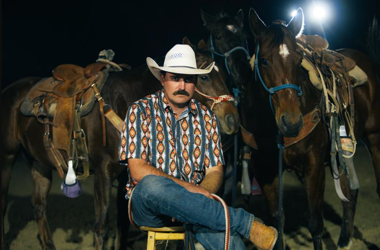 Country music artist Wade Forster in a cowboy hat, mustache, and patterned shirt sitting in front of saddled horses at night under stage lights.